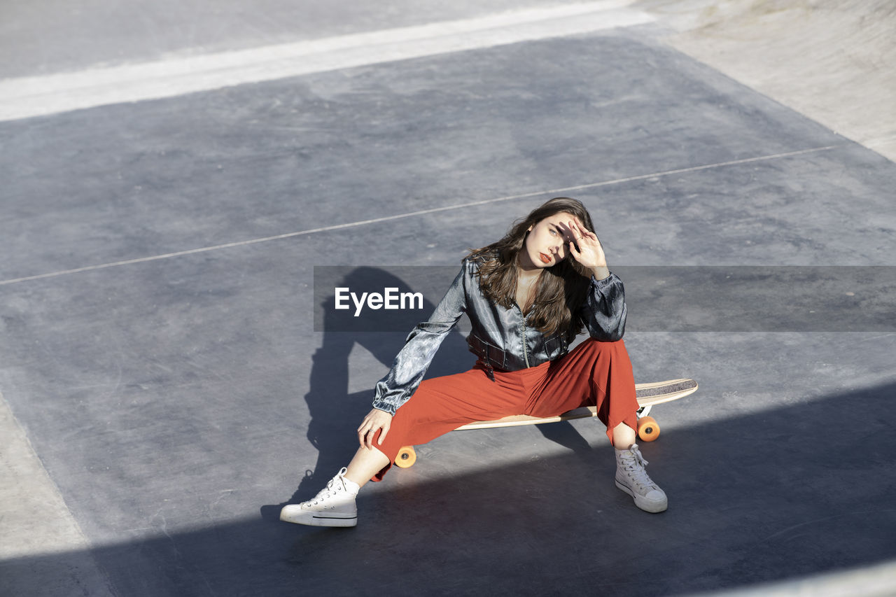 From above of young woman in stylish clothes touching forehead while sitting on longboard and looking at camera