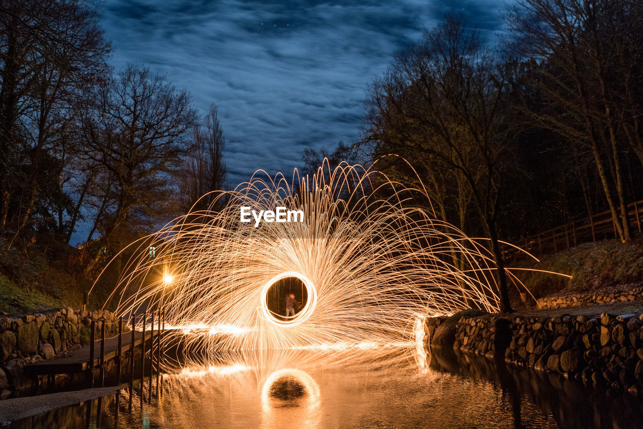 Blurred motion of illuminated trees by river against sky at night