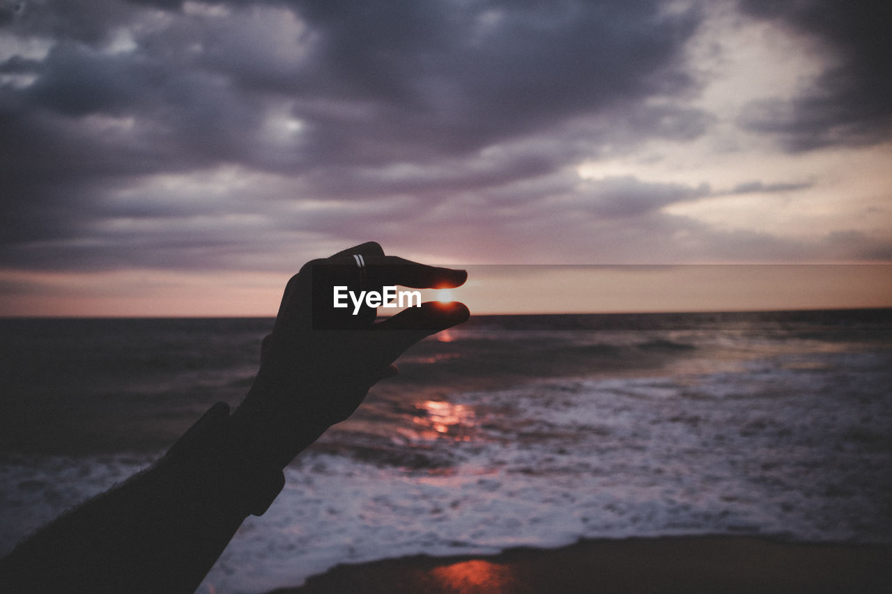 Optical illusion of silhouette hand holding sun against sea and sky during sunset