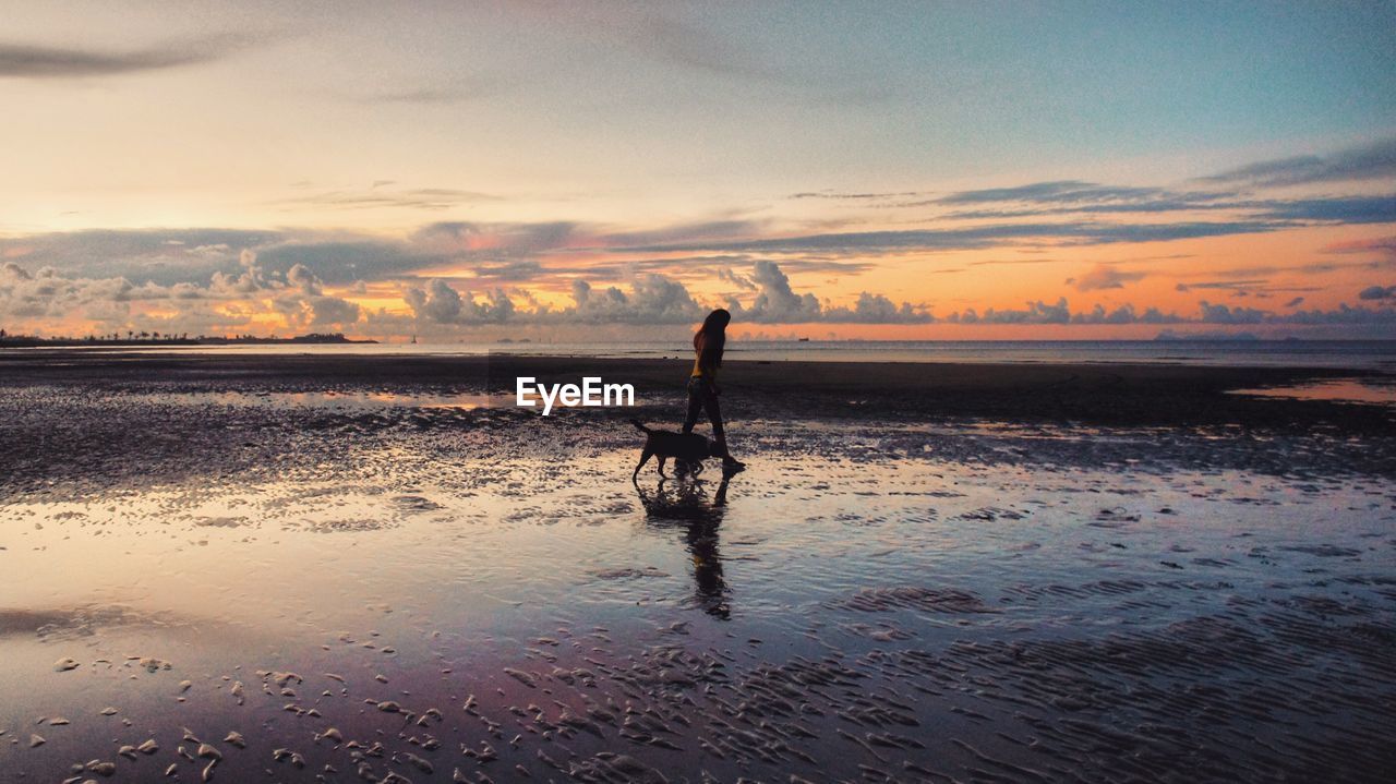 Woman walking on shore at beach against sky during sunset