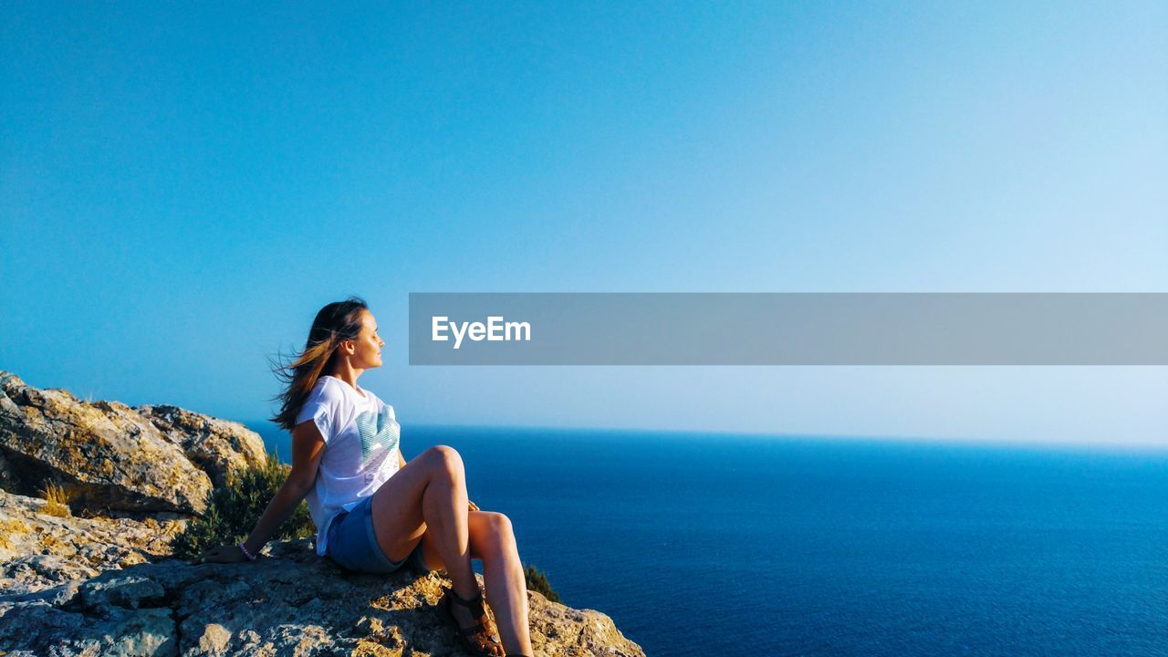 Woman sitting on rock by sea against clear blue sky