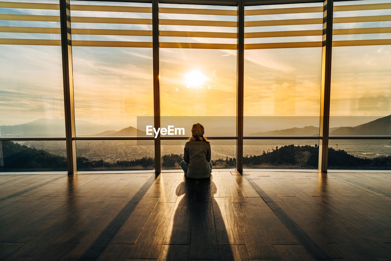 Rear view of young woman looking through window during sunset