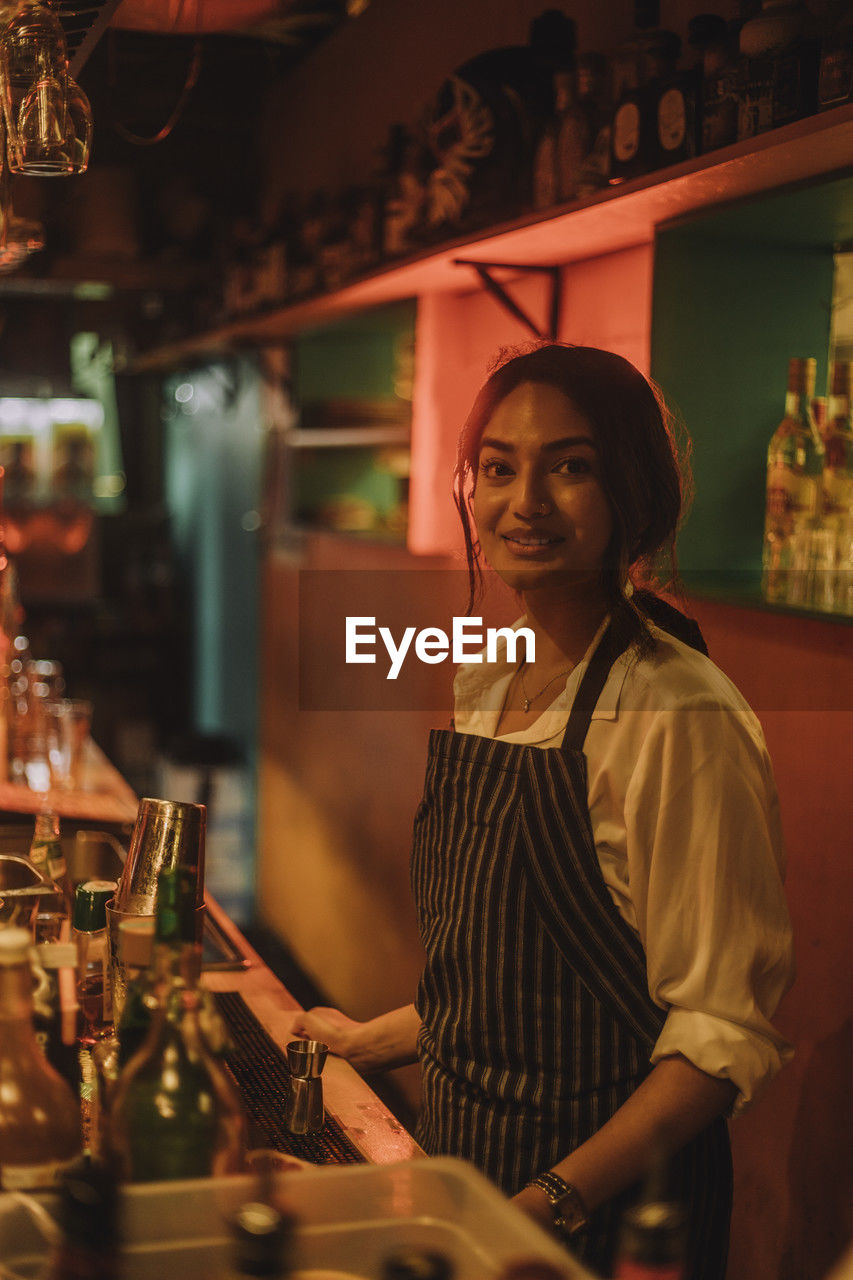 Portrait of smiling female bartender wearing apron and standing at bar