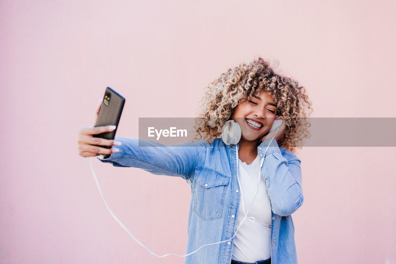 Portrait of smiling hispanic woman with afro hair in city using mobile phone and headset. lifestyle