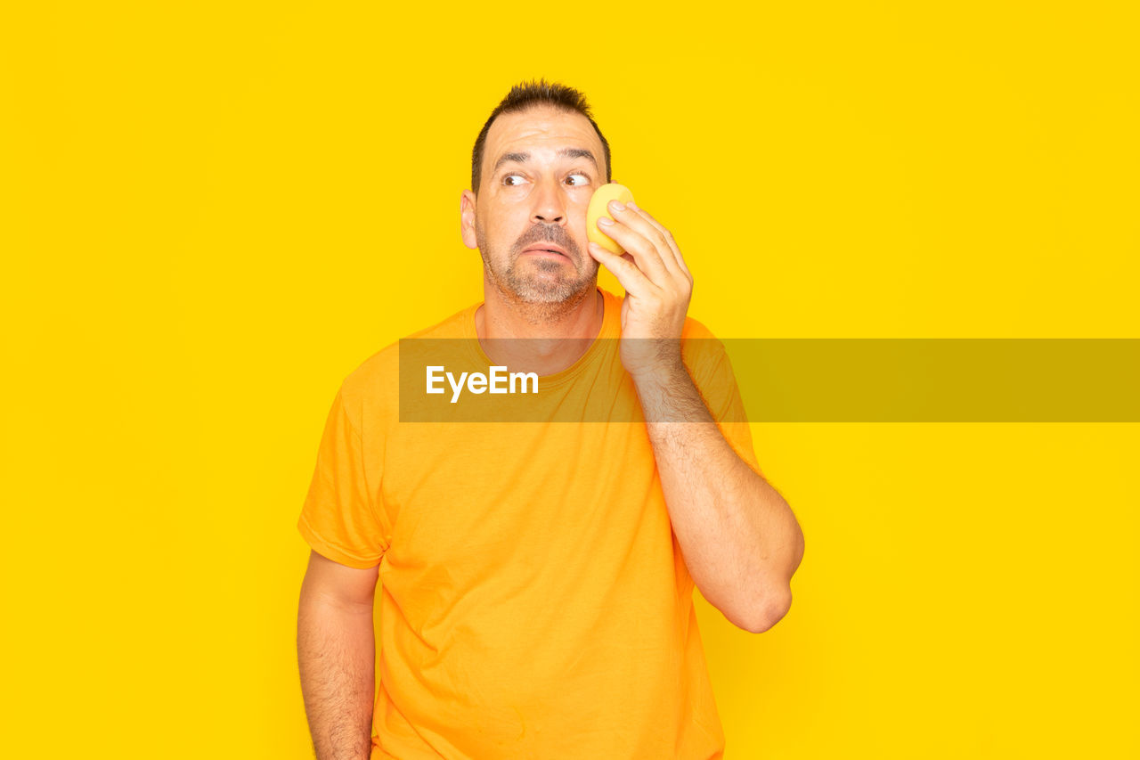 yellow, adult, one person, studio shot, yellow background, colored background, men, indoors, person, orange, orange color, waist up, portrait, finger, human face, casual clothing, copy space, hand, front view, clothing, facial hair, standing, holding, looking at camera, beard, t-shirt, food and drink, emotion, lifestyles, humor, fun