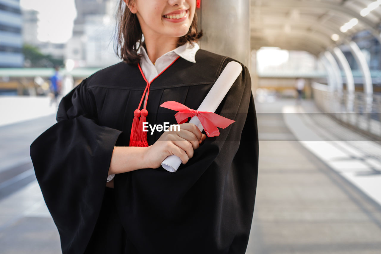 Midsection of woman wearing graduation gown while holding certificate against in city