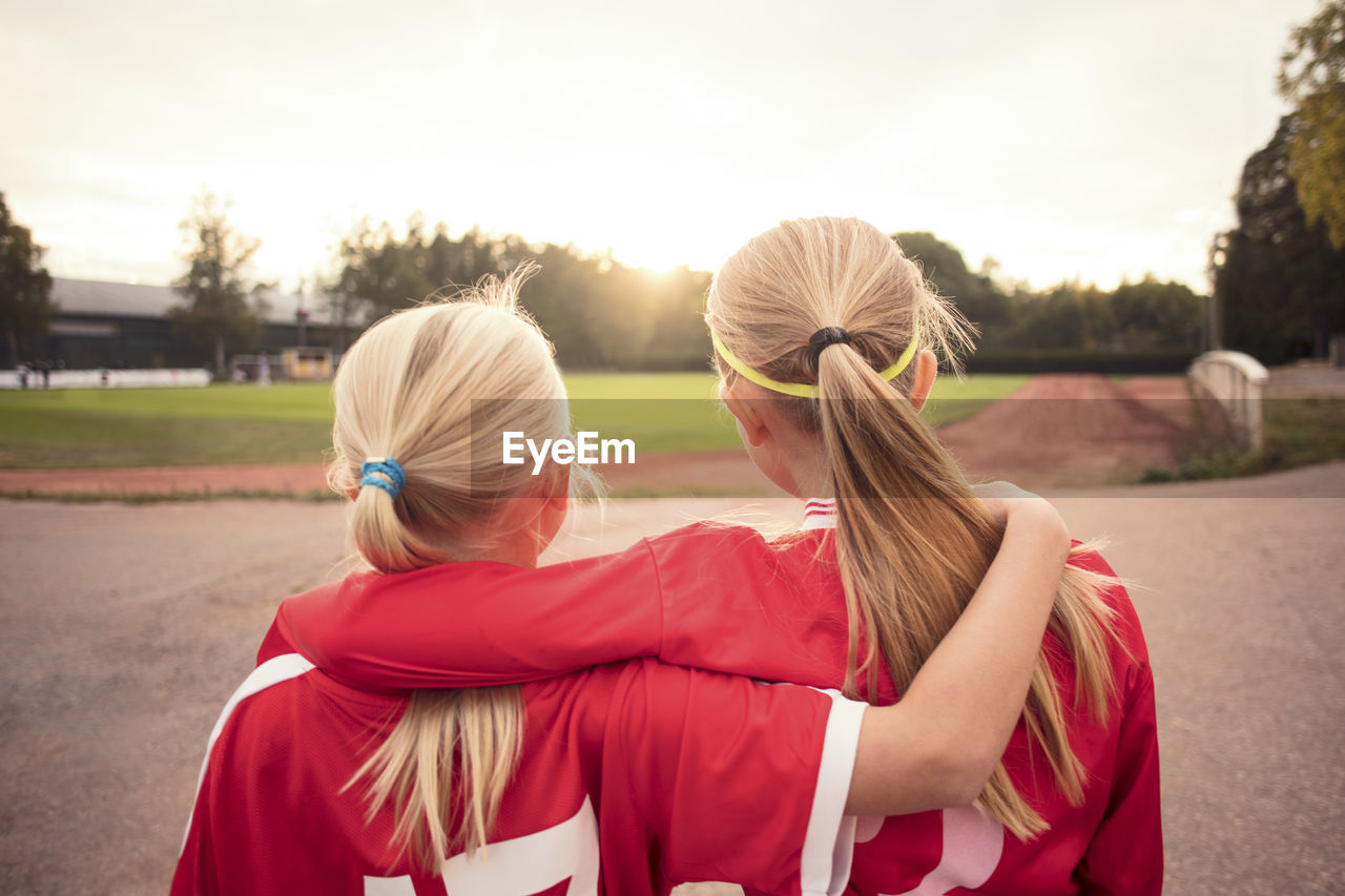 Rear view of female soccer friends standing on footpath looking at field during sunset