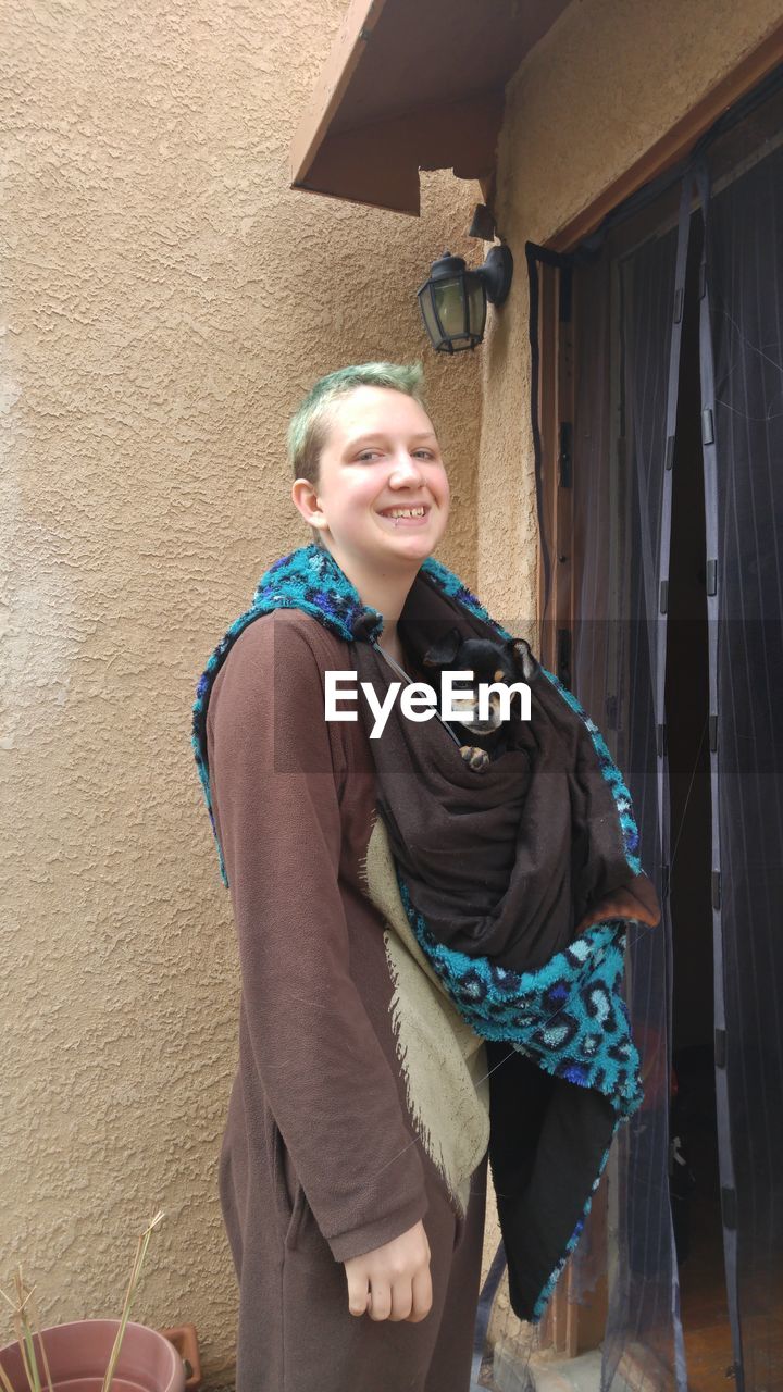 Portrait of smiling woman carrying dog in blanket while standing by house