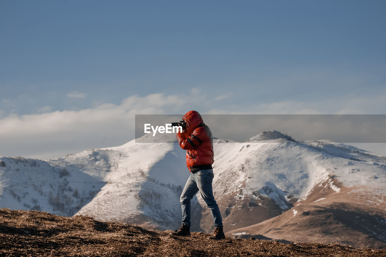 Man with camera taking photos on snowcapped mountain against sky