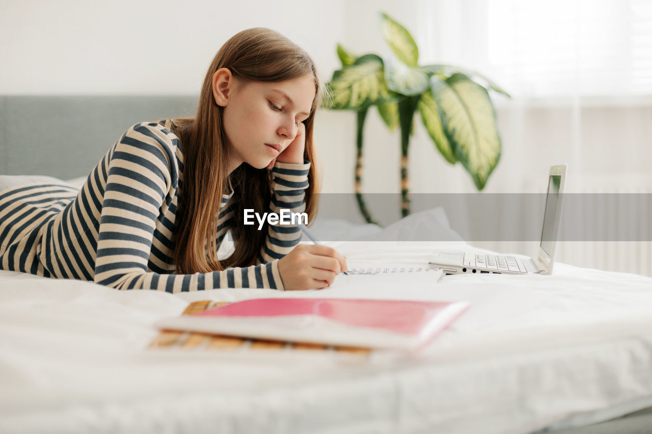 A charming teenage girl is lying on the bed, doing homework with a laptop, making notes 