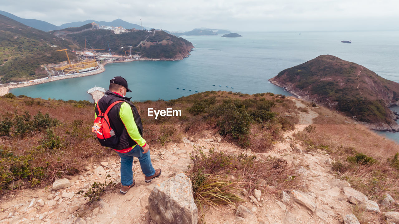 MAN STANDING ON MOUNTAIN BY SEA AGAINST MOUNTAINS