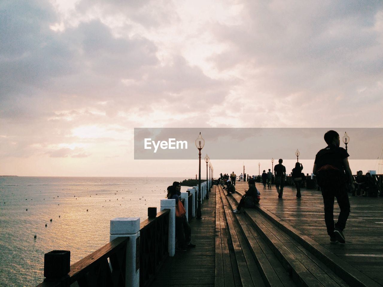 People on pier over sea against cloudy sky during sunset