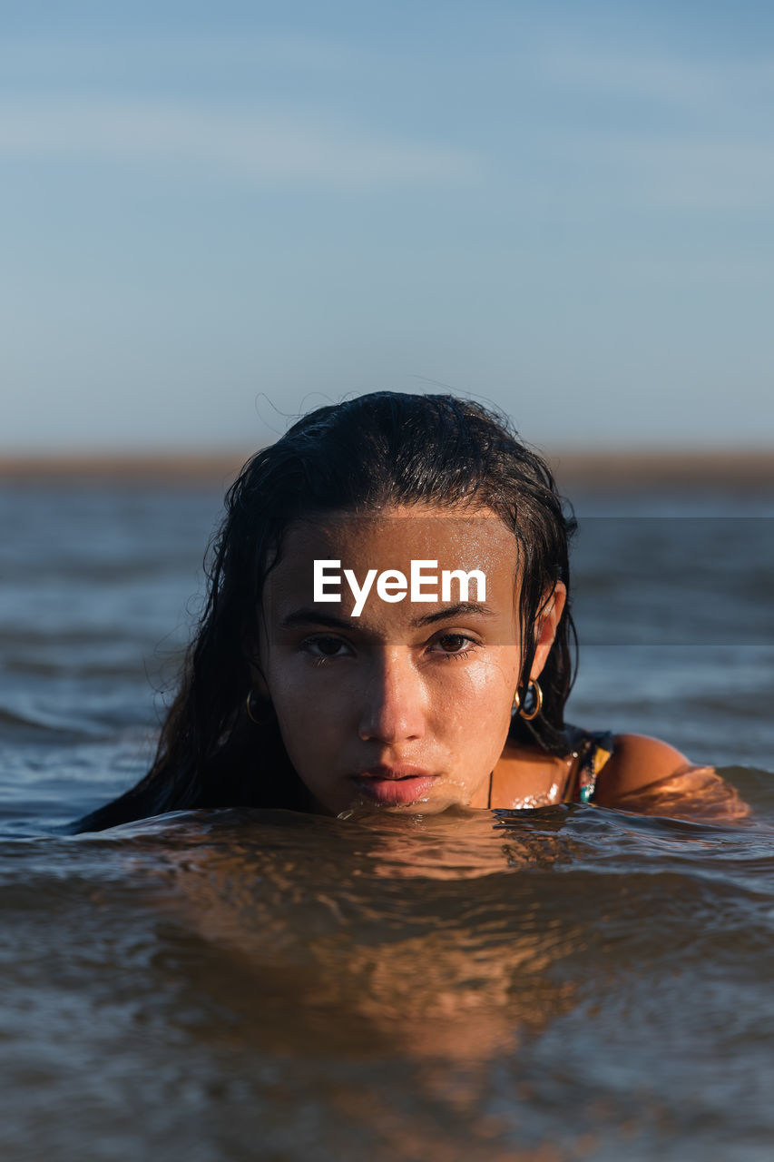 Serene female with wet hair swimming in calm sea in summer evening and looking at camera