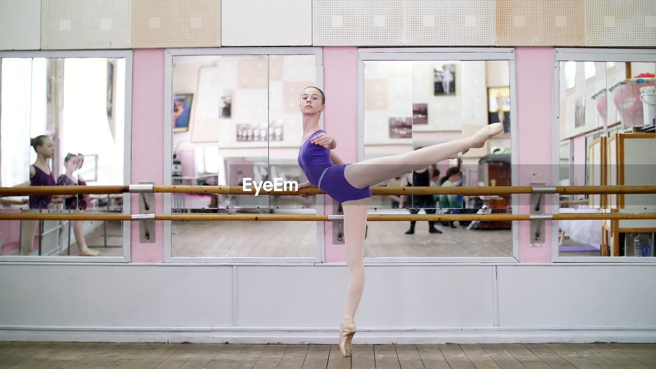 In dancing hall, young ballerina in purple leotard performs developpe attitude on pointe shoes