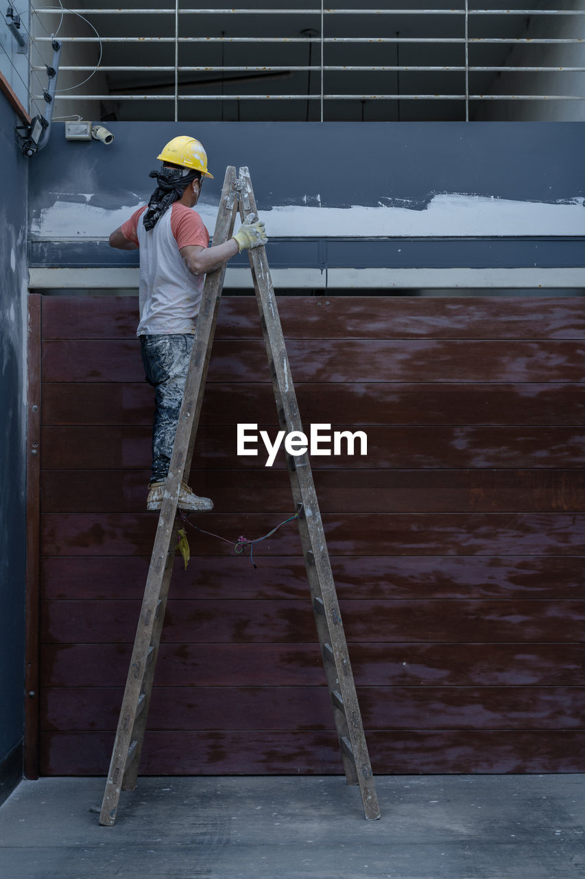 Side view of professional male painter in yellow hardhat in process of plastering wall while standing on a ladder on sidewalk on city street