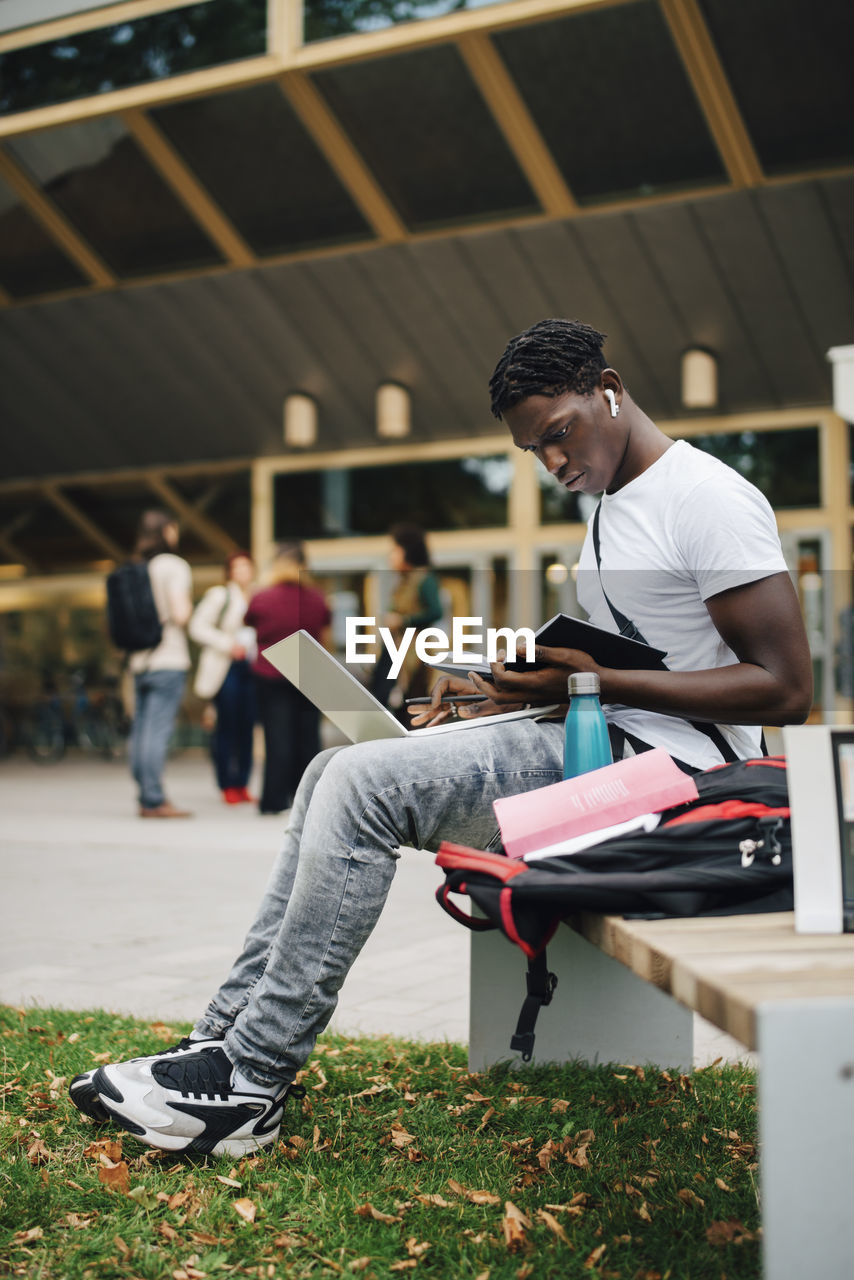 African male student reading book while sitting on bench in university campus