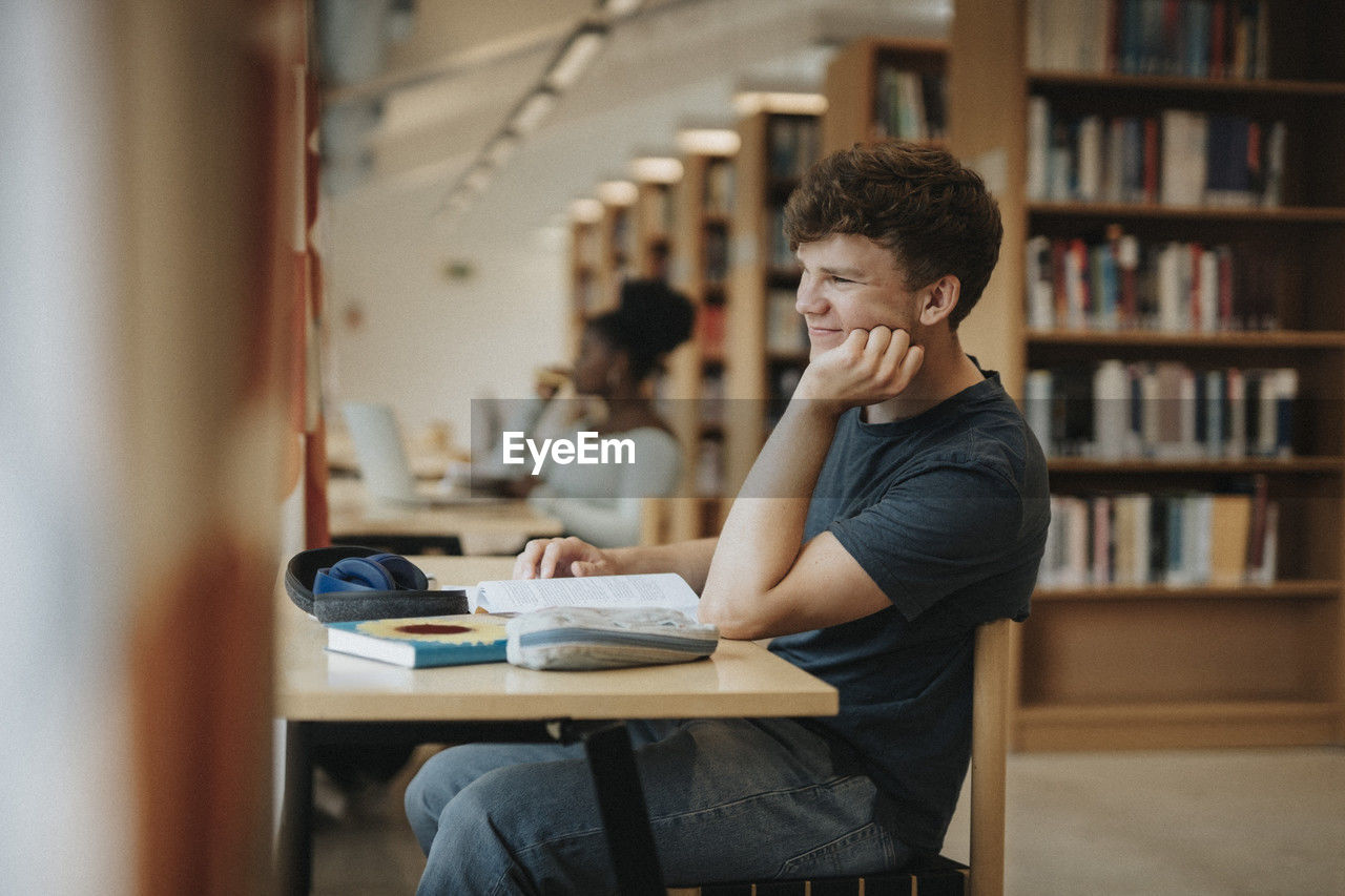 Side view of smiling student sitting with hand on chin in library