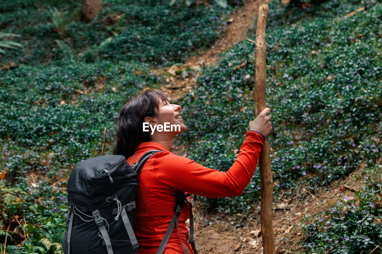 Side view of glad woman with backpack and stick admiring forest while travelling through coniferous woodland in weekend