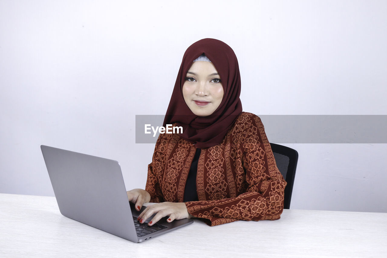 PORTRAIT OF YOUNG WOMAN USING SMART PHONE AGAINST WALL