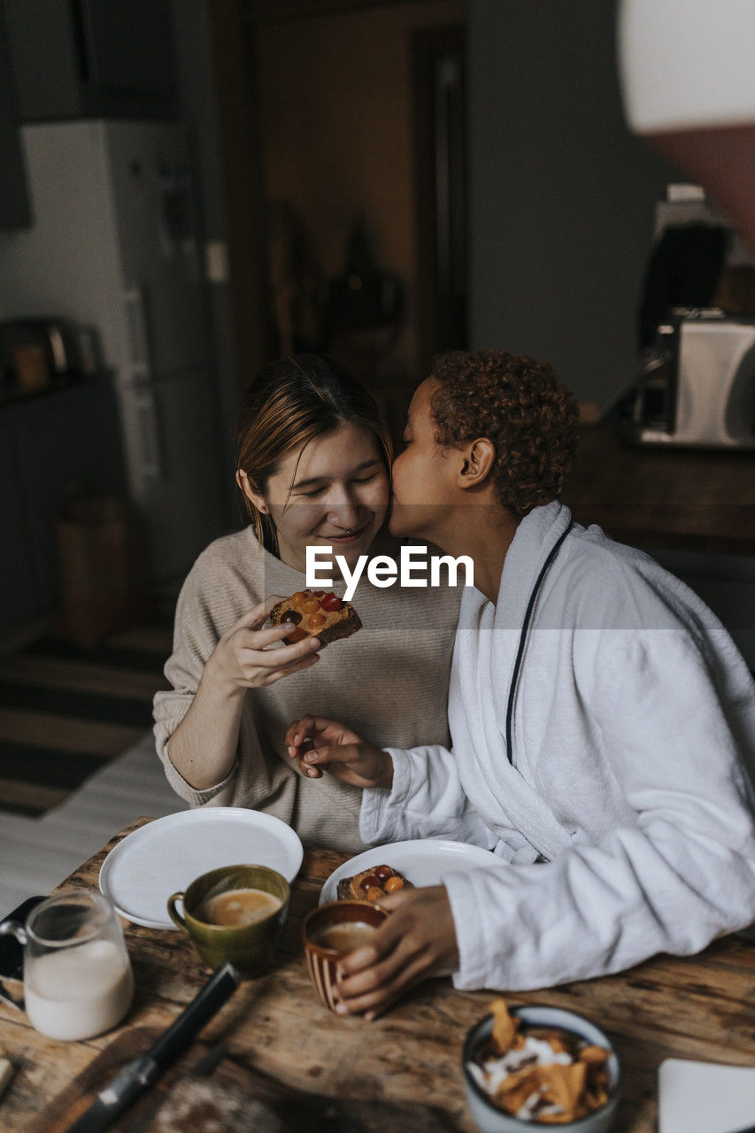 Non-binary person kissing friend while having breakfast at home