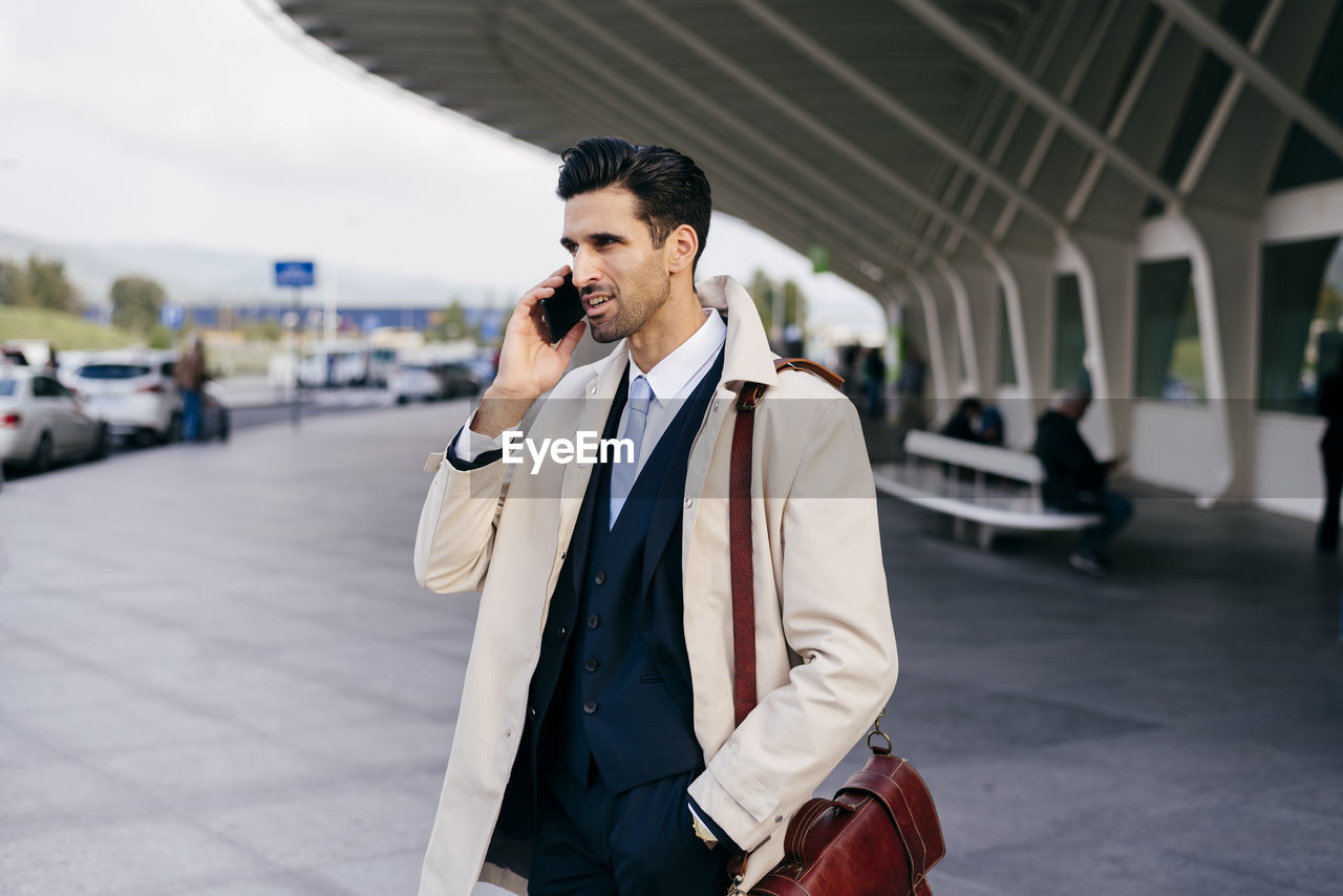 Busy man with dark hair in suit and coat speaking on phone while standing near airport terminal at daytime