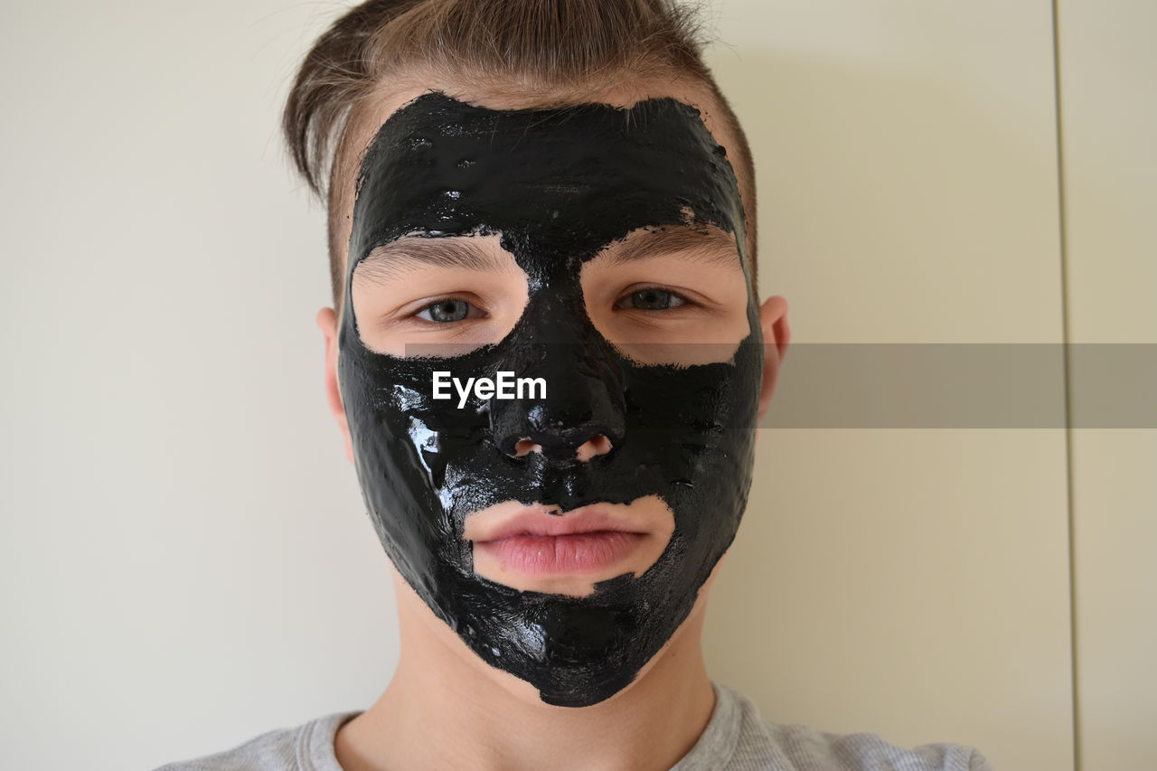 Close-up portrait of teenage boy with black facial mask against wall