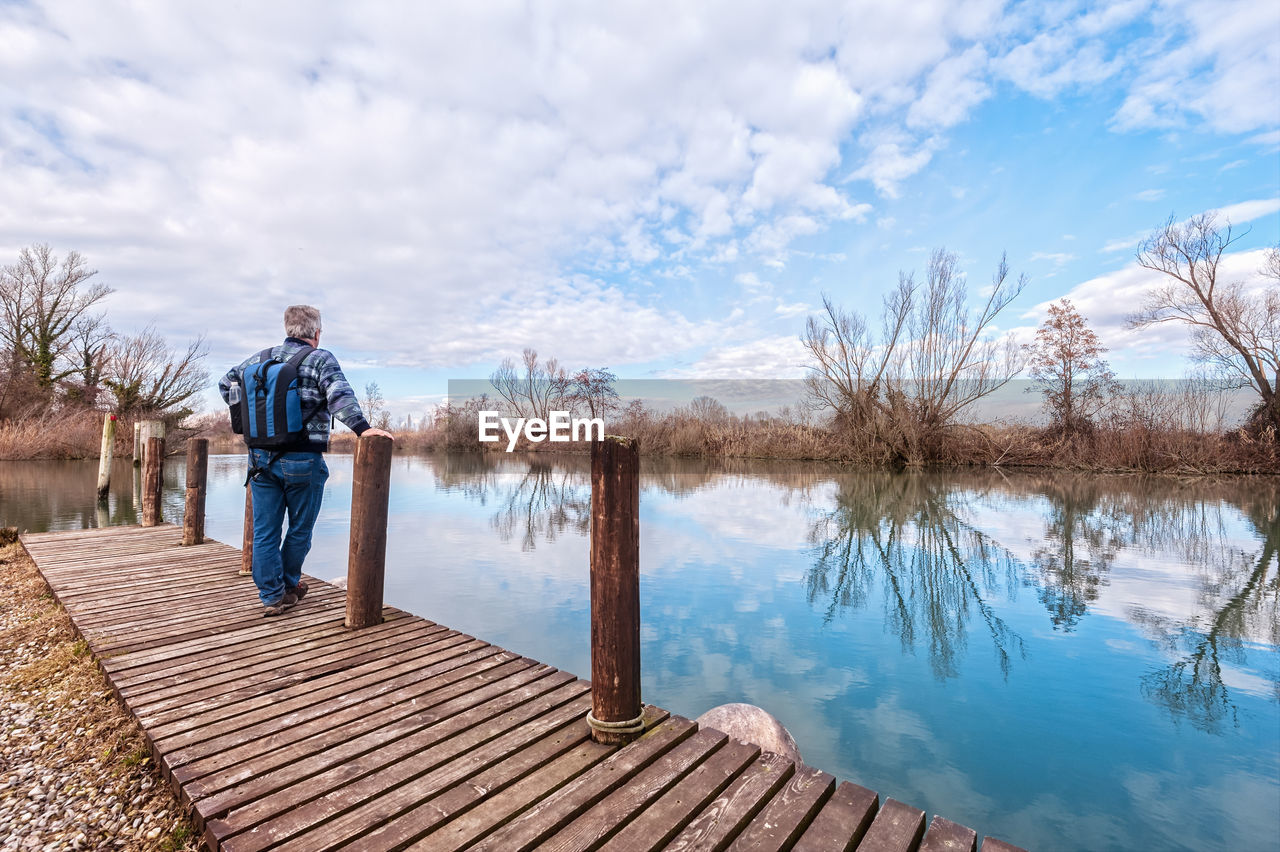 Rear view of mature man with backpack standing on pier over lake against cloudy sky