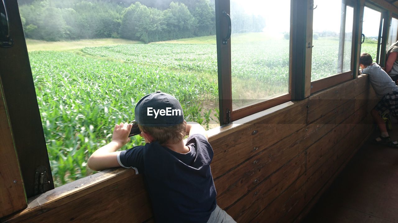 Rear view of boys leaning on window against field