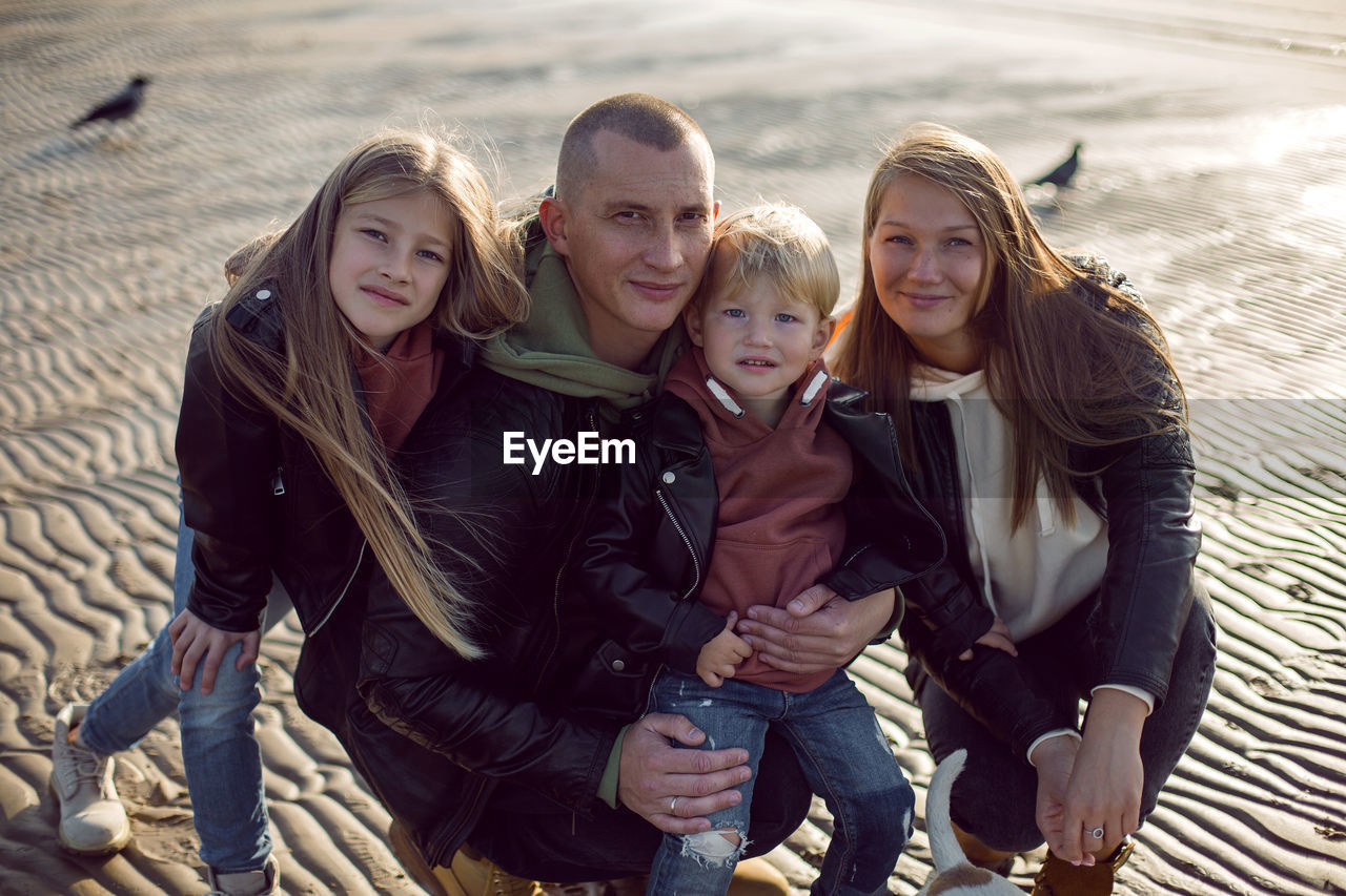 Family in a leather jacket stands along the beach with their dog in autumn