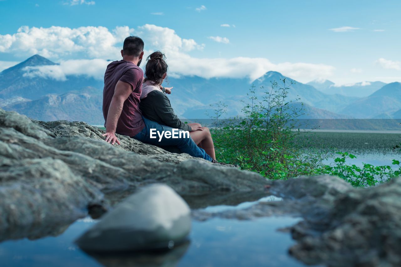 Friends sitting on rock by lake against sky