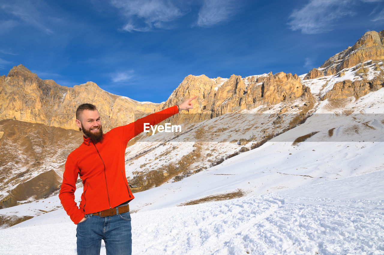 A bearded attractive caucasian man in a red sweater and jeans points with his hand to the mountains 