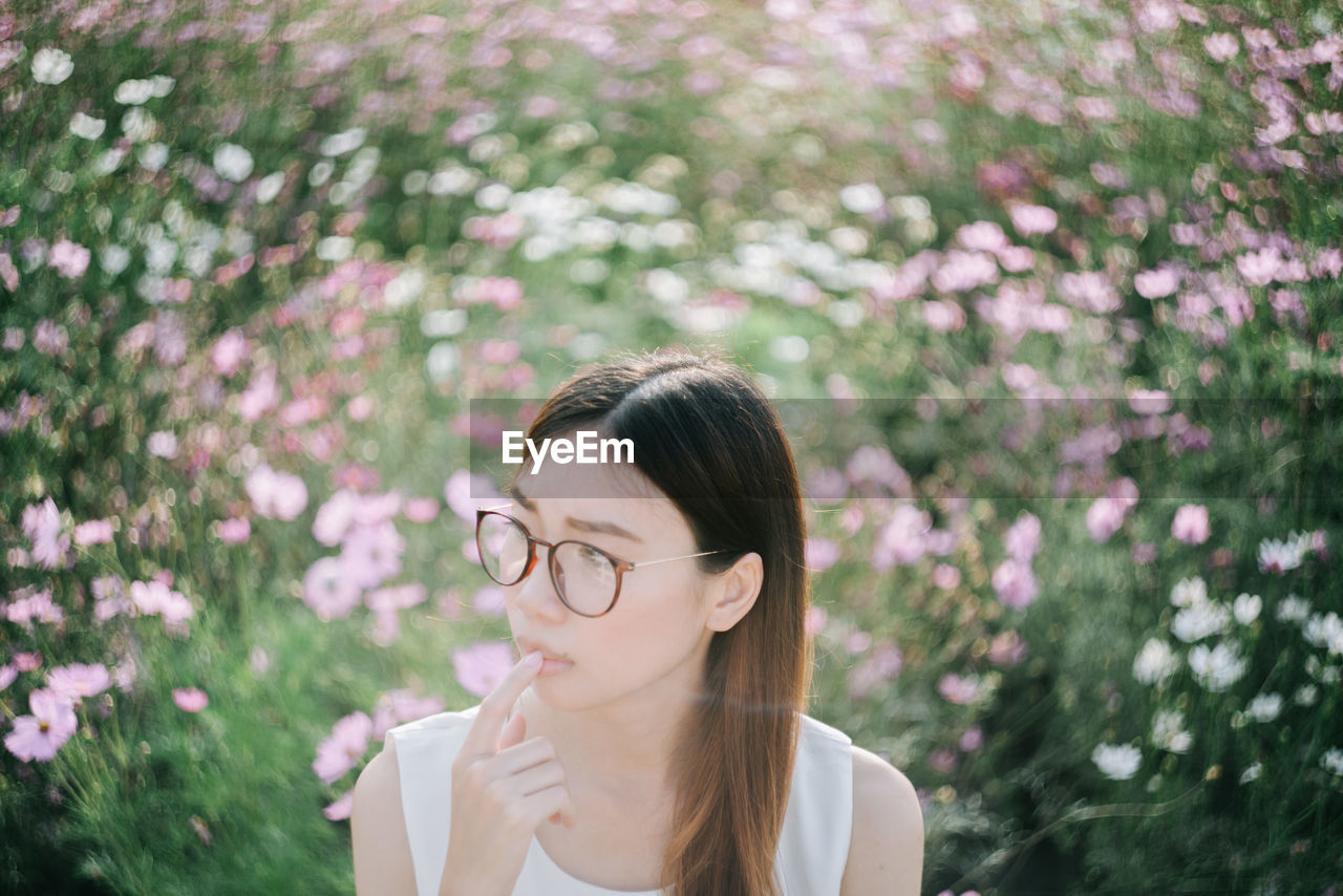 Thoughtful young woman wearing eyeglasses looking away against flowers