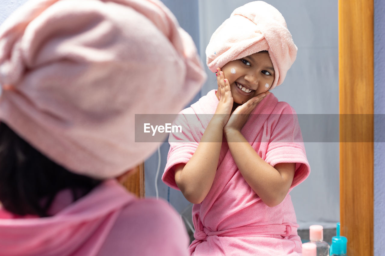 A cute indian girl child in pink bathrobe applying face cream and touching cheeks in front of mirror