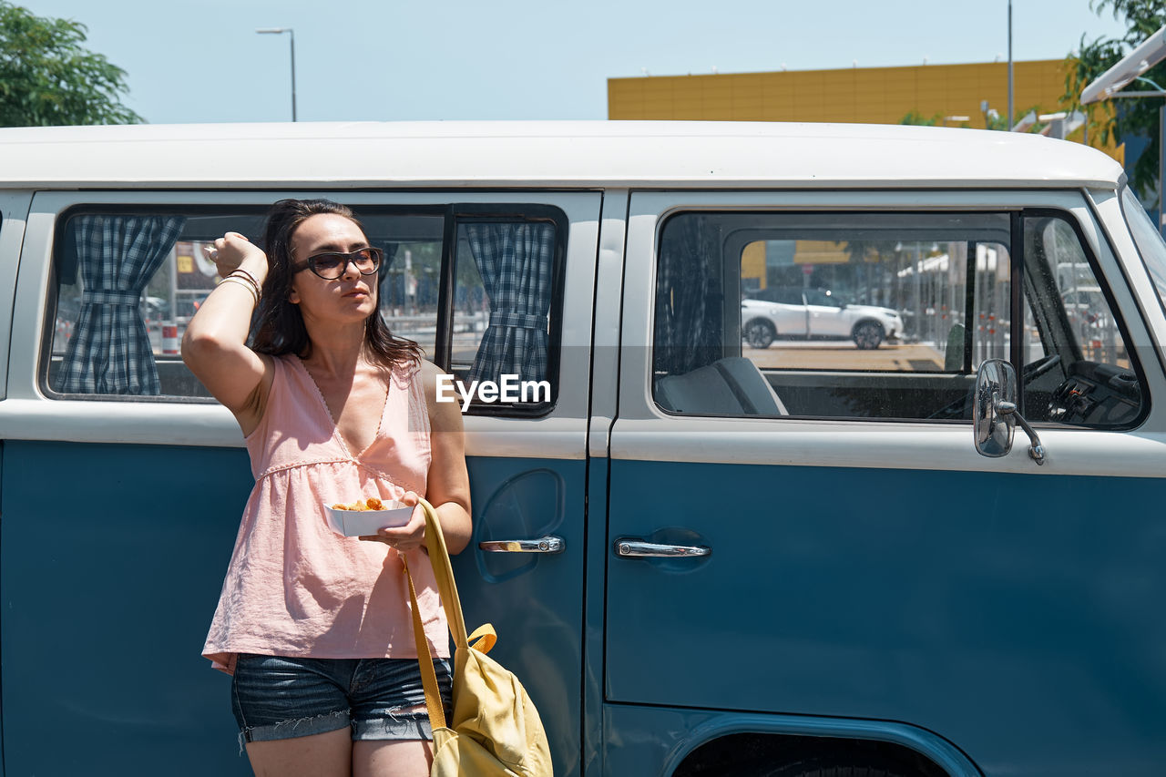 Young woman eating unhealthy fast food near a camper van in urban parking. traveling and summertime. 