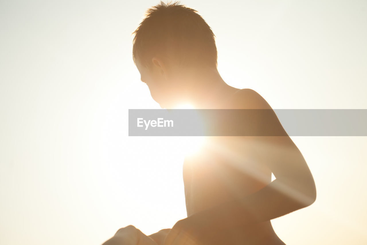 Silhouette shirtless boy against bright sky