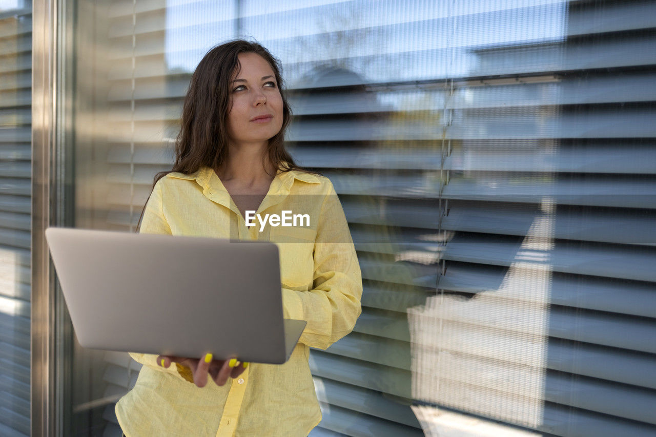 Thoughtful businesswoman standing with laptop near window