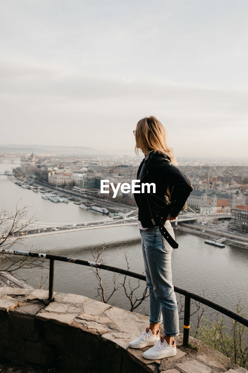 Young woman standing on retaining wall against cityscape during sunset