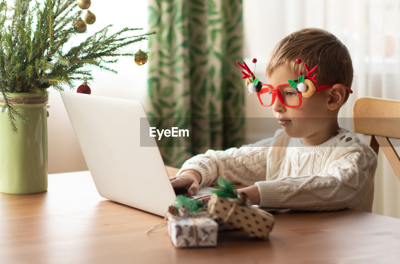 Little boy in a white sweater and funny glasses sits at a laptop and writes a letter to santa claus