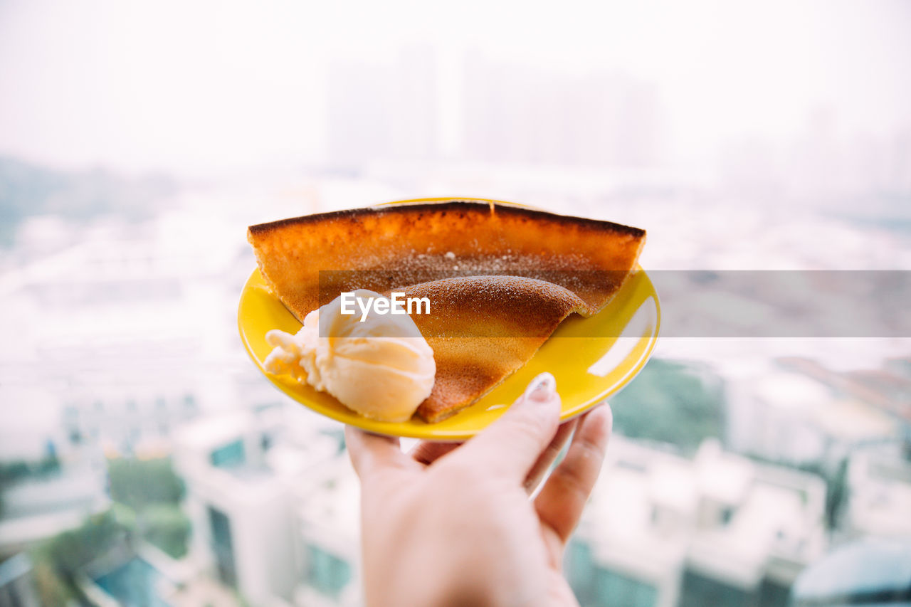 Cropped image of woman hand holding ice cream with pancake in plate against cityscape