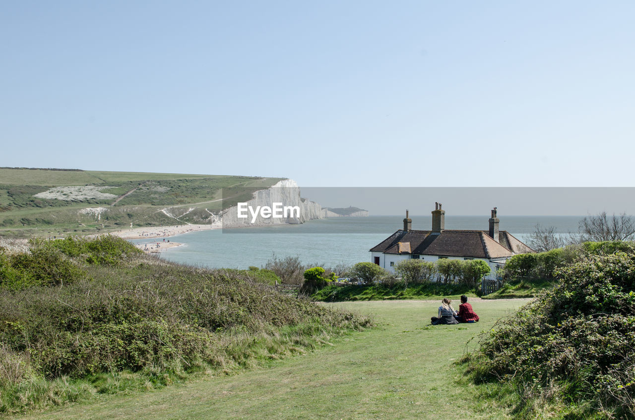 Two people staring at scenic view of field against clear sky with a beach house in the background 