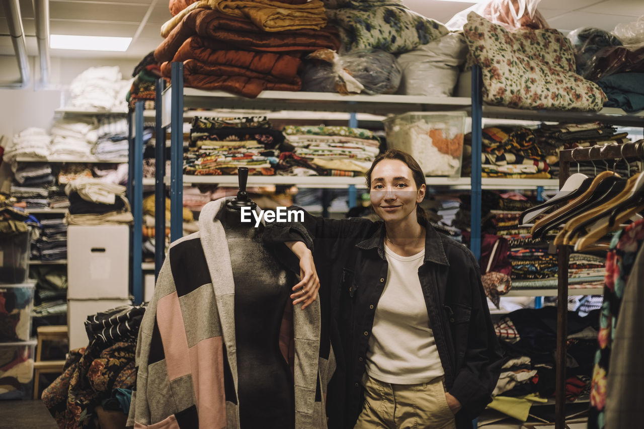 Portrait of female fashion designer with hand in pocket leaning on mannequin at workshop