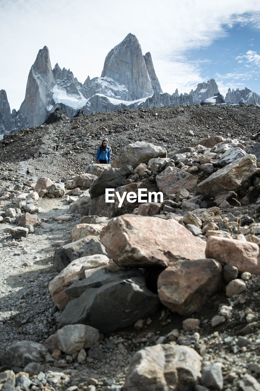 Woman on rocks by snowcapped mountains against cloudy sky
