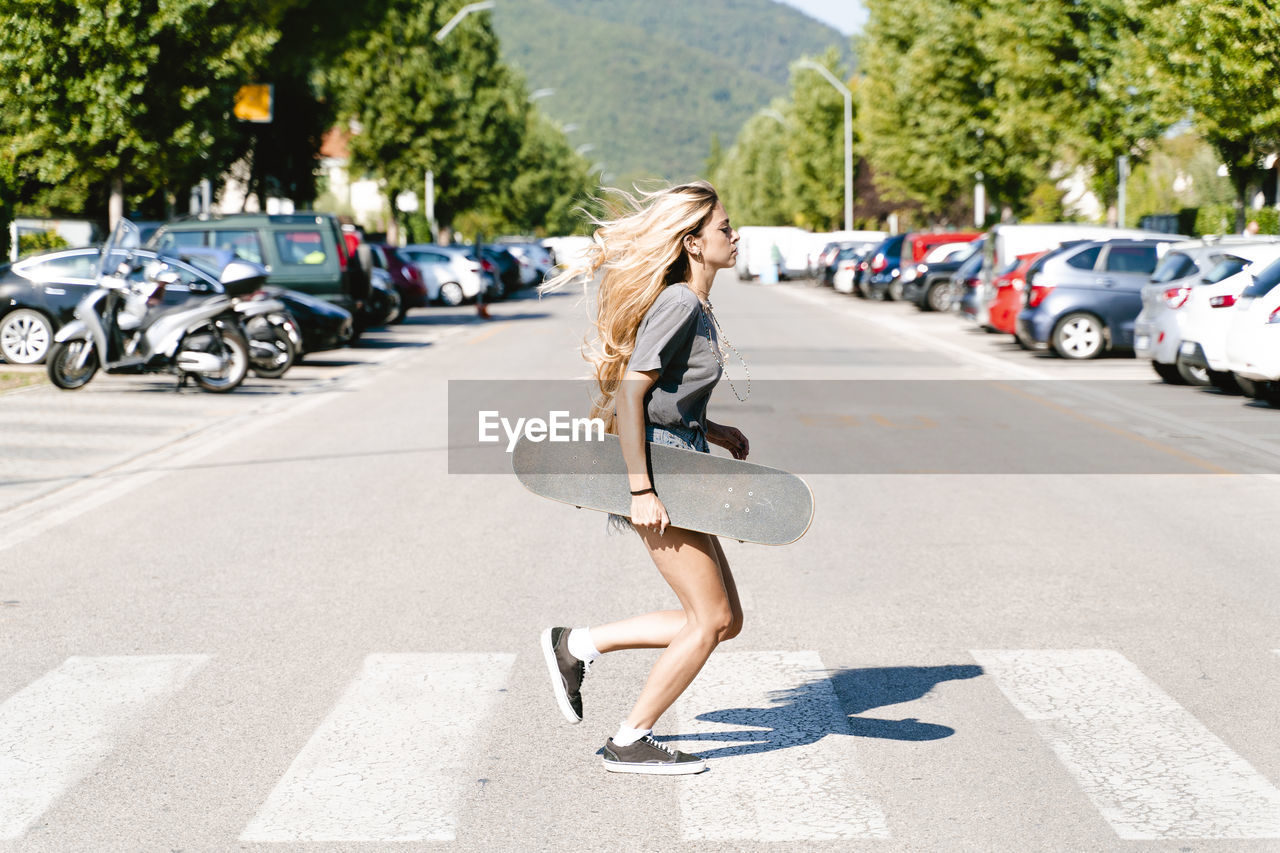 Young blond woman holding skateboard while running on zebra crossing in city
