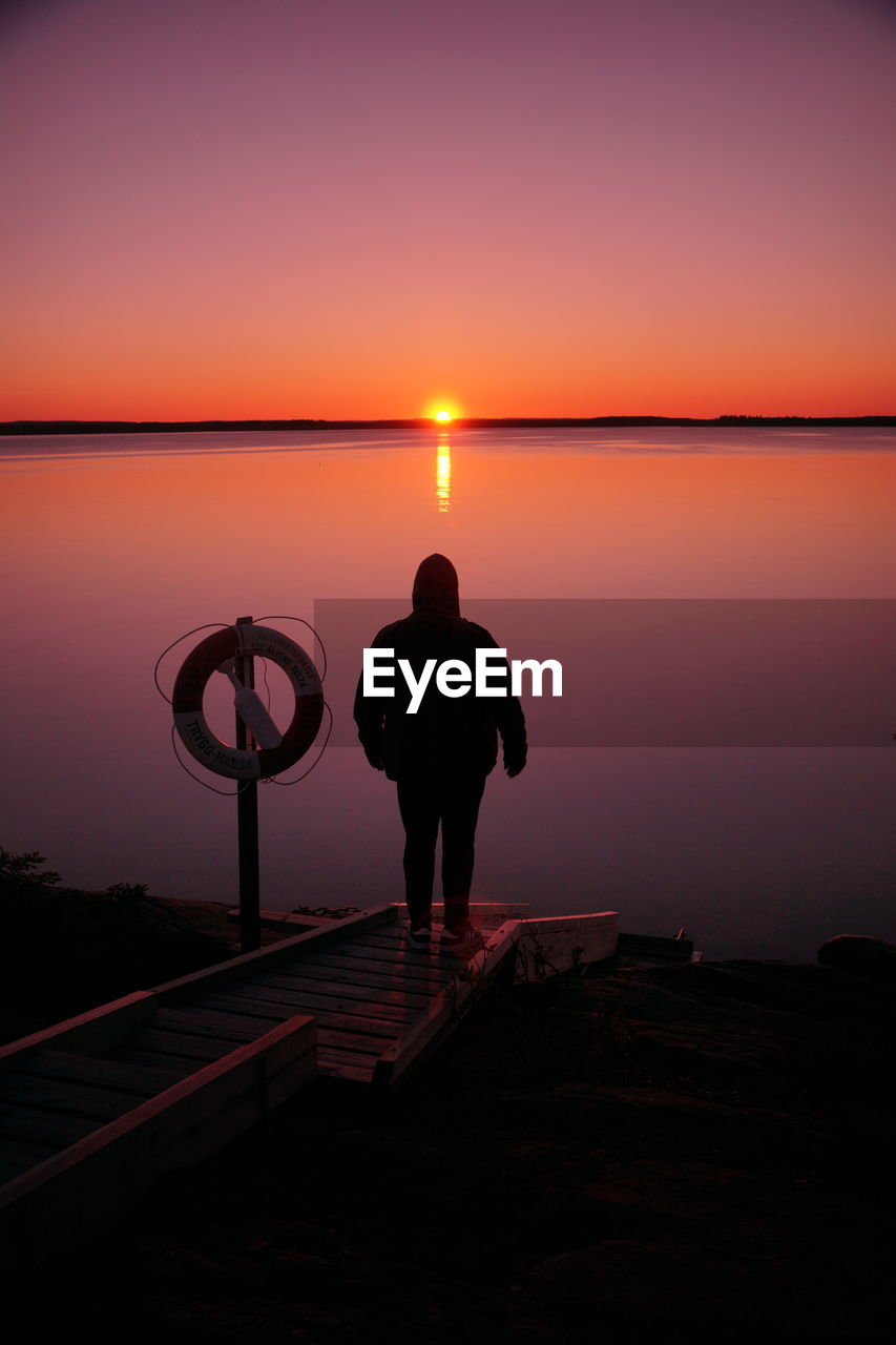 Rear view of silhouette man standing on shore against sky during sunset