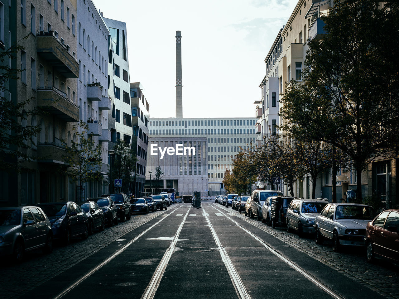 Empty road amidst cars parked by buildings against sky