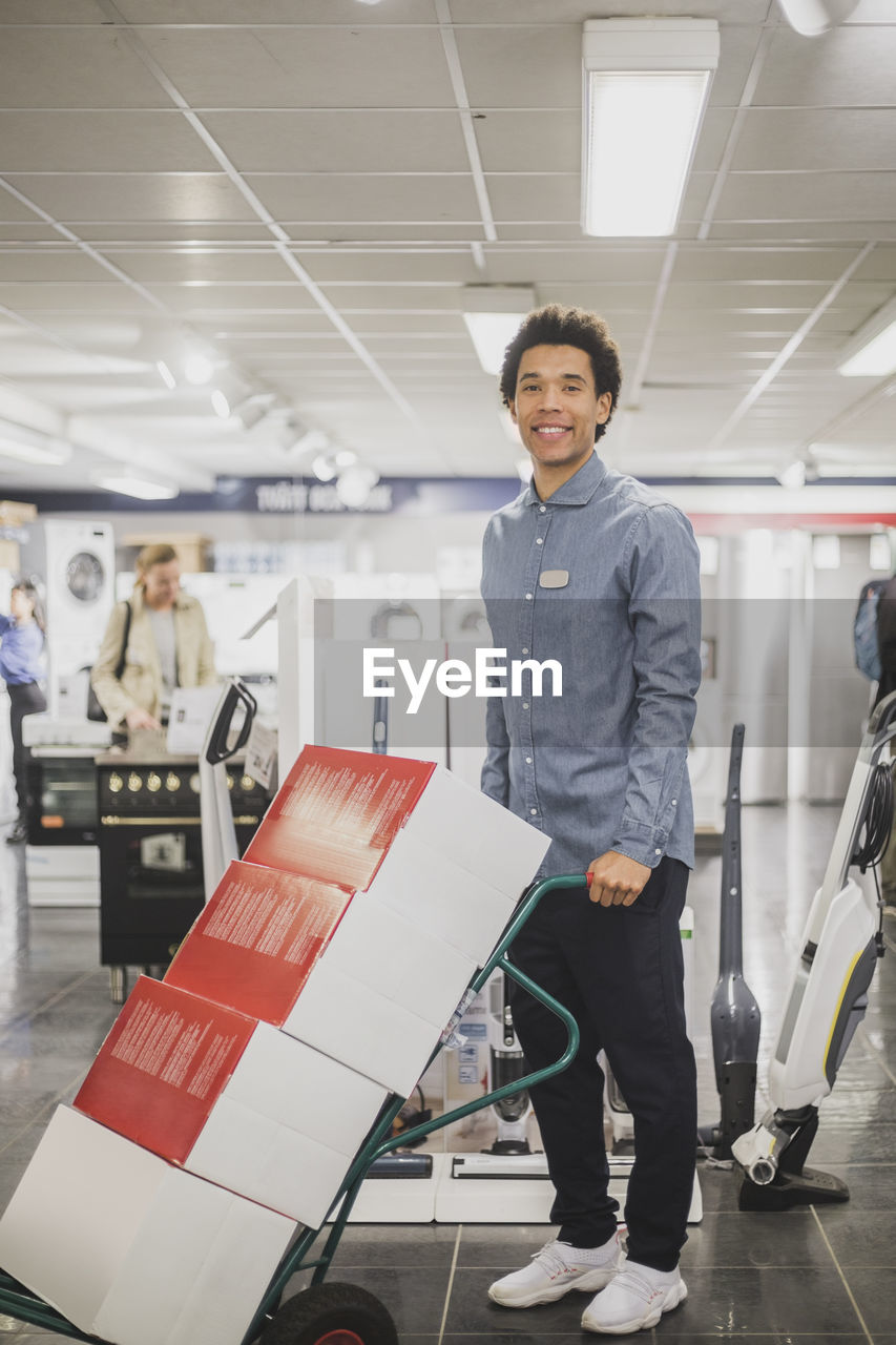 Portrait of smiling male owner with luggage cart standing in electronics store