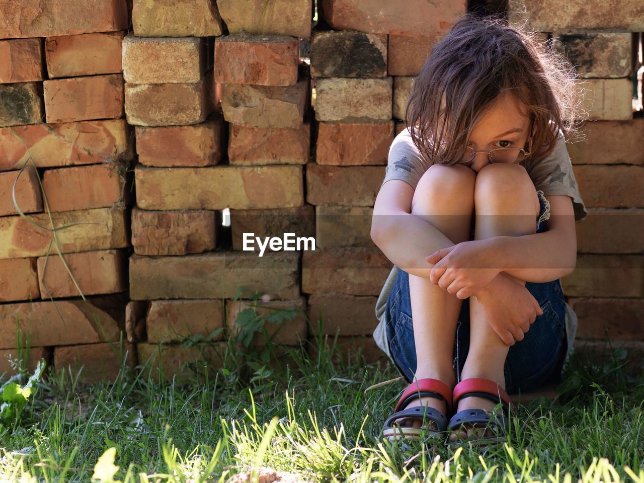 Depressed boy hugging knees while sitting on grass against brick wall