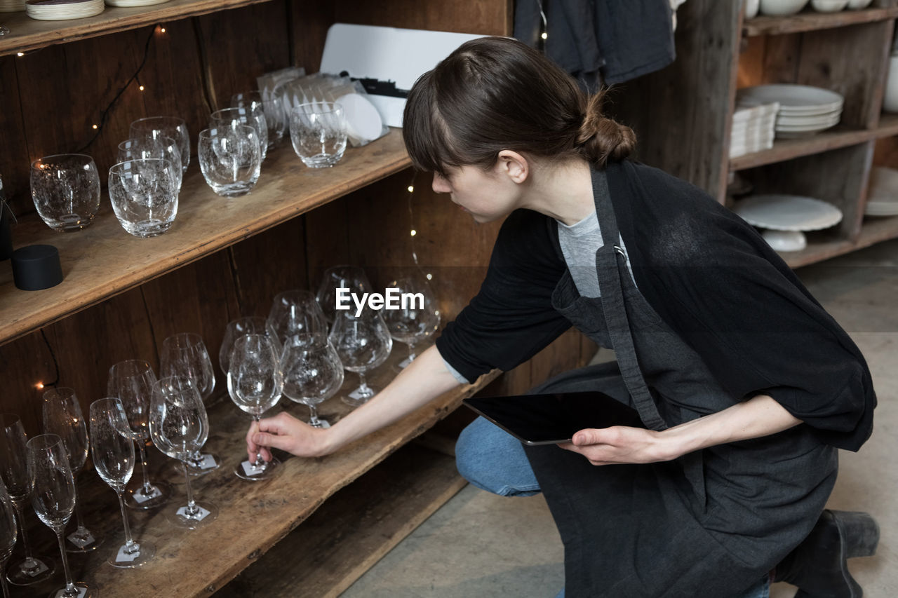 High angle view of young female owner with tablet arranging wineglass while crouching by rack at store