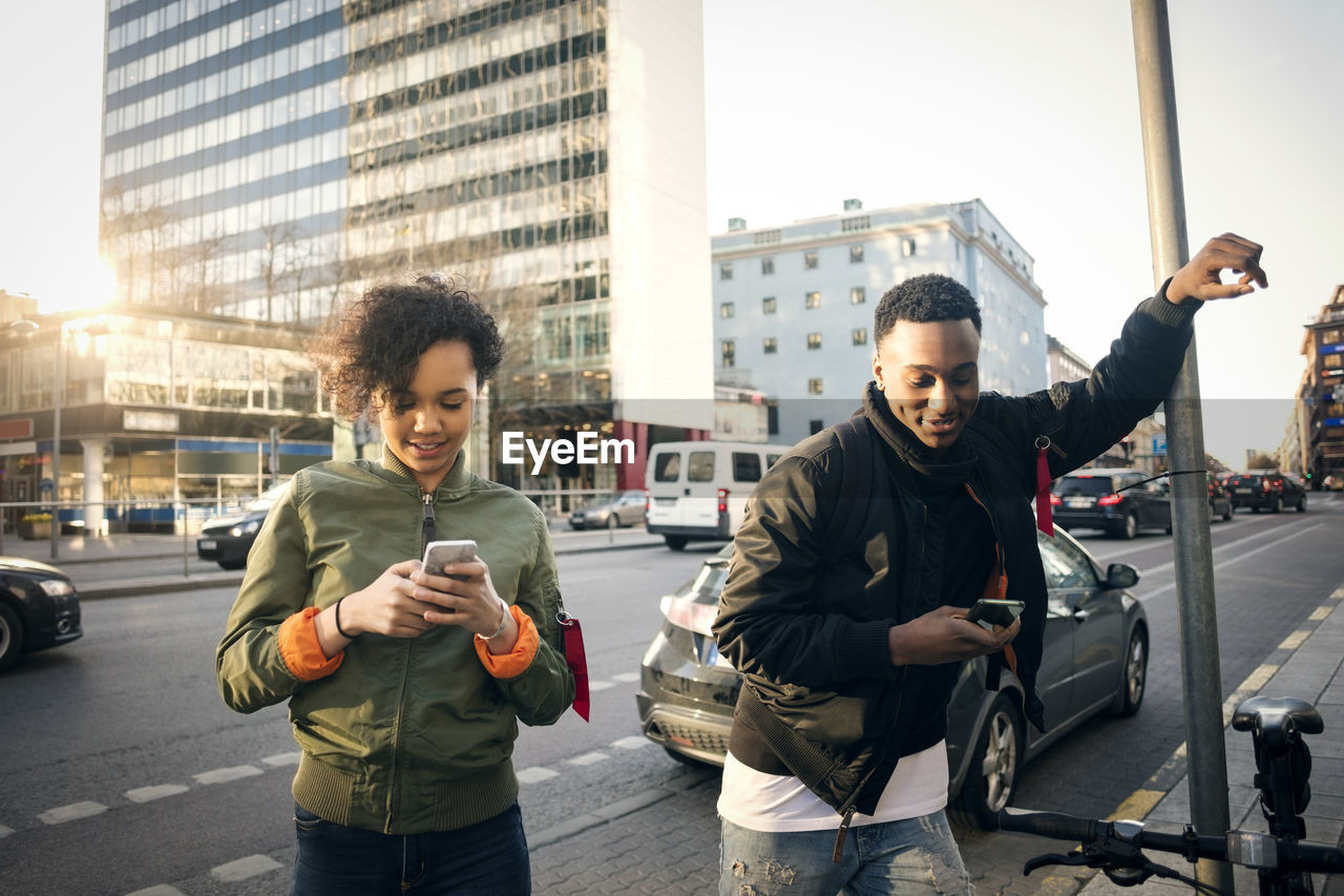 Teenagers using phone while standing on sidewalk by city street