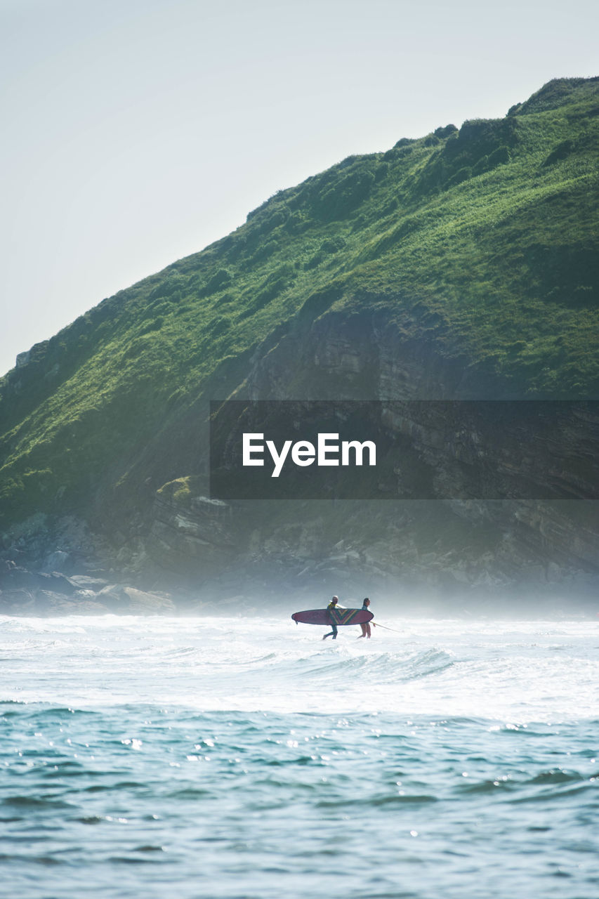 Man and woman holding surfboard in sea against mountain